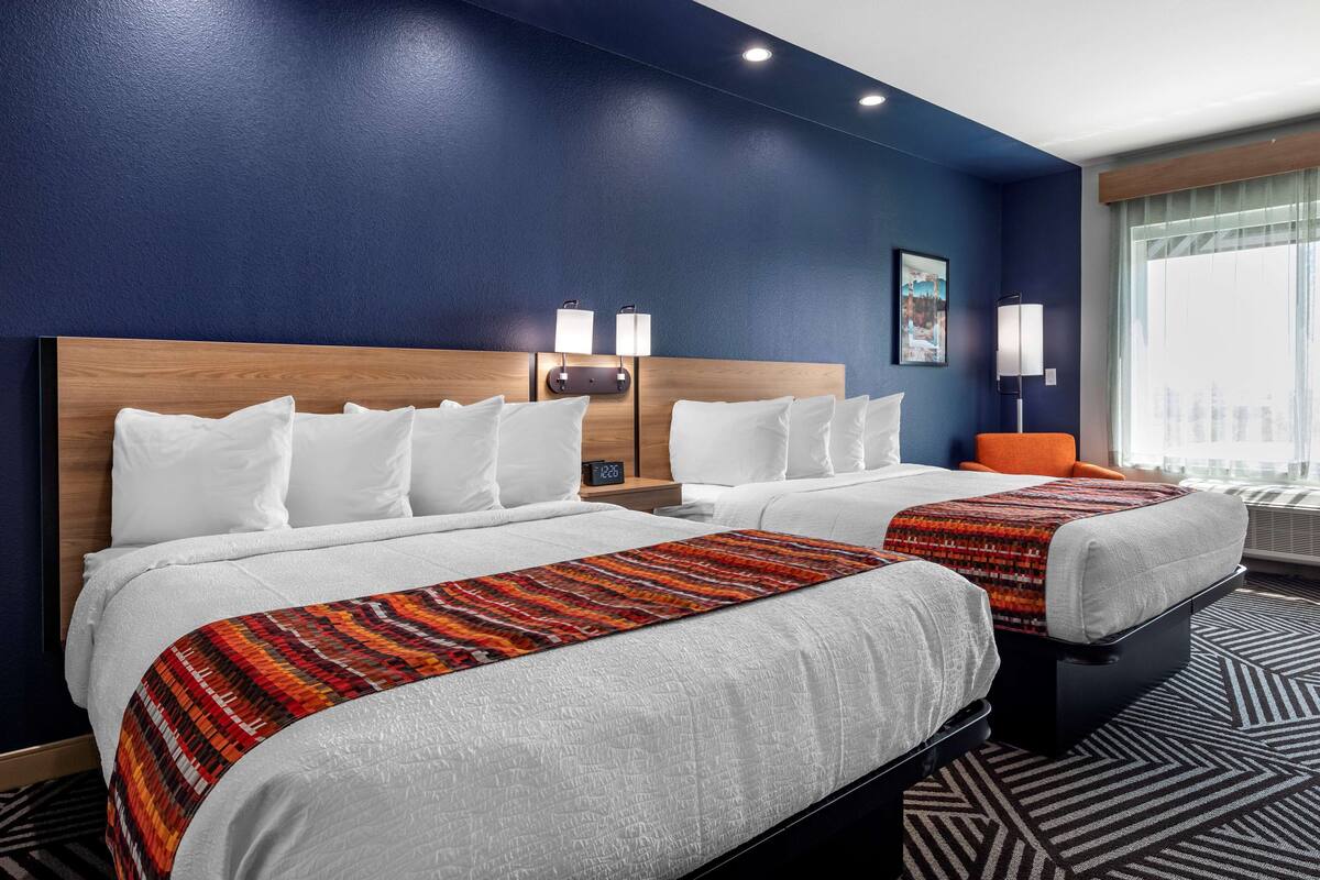 Een hotelkamer met twee bedden, een nachtkastje met lamp, een raam met gordijnen en een kamerbreed tapijt met dessin.
