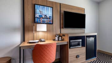 Habitación de hotel con un escritorio de madera, microondas, refrigerador, televisor y un cuadro de un paisaje urbano.