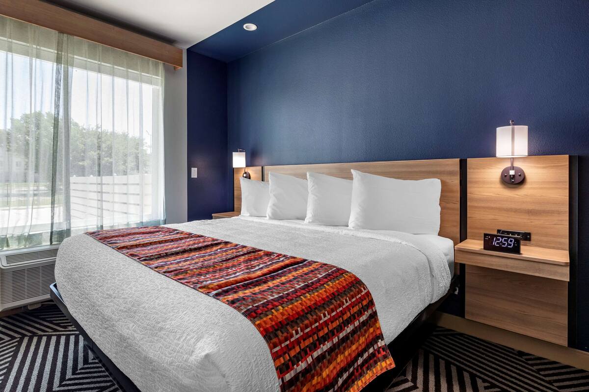 Een hotelkamer met een groot bed, een nachtkastje met digitale klok en een raam met dunne gordijnen.
