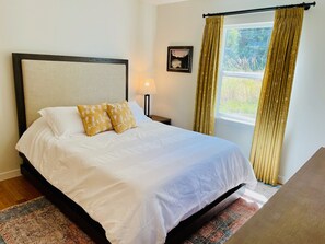 A neatly made bed with white linens and two decorative pillows, a bedside lamp, a framed picture on the wall, and a window with curtains.