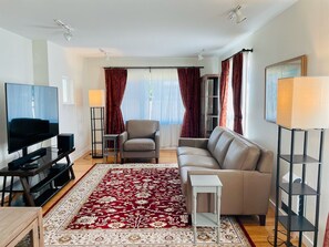 A modern living room with a flat-screen TV, a grey sofa, a red patterned rug, and a small white table.
