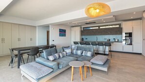 Een moderne woonkamer met een hoekbank, een eettafel met stoelen, een keuken met kastjes en apparatuur, en een hanglamp.
