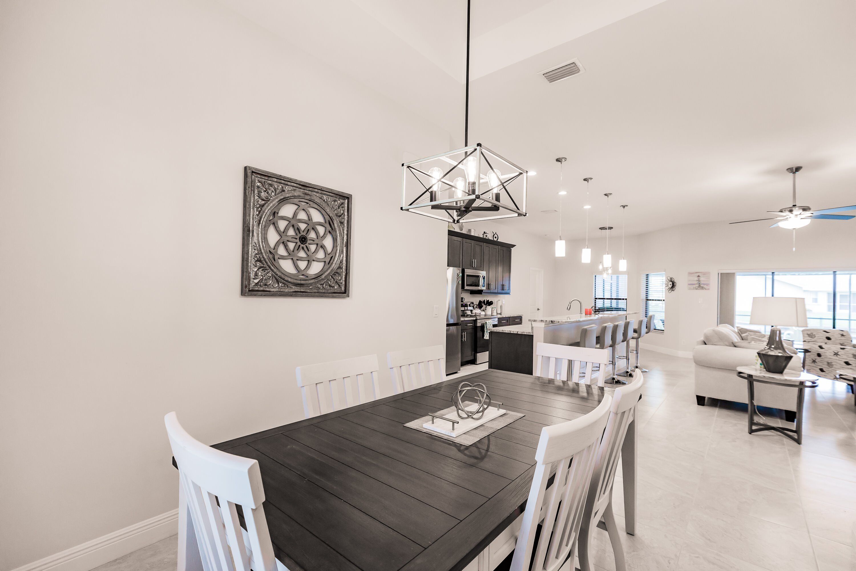 A modern kitchen with dark countertops, white cabinetry, and a central island. There is a dining area with a dark wooden table and white chairs.