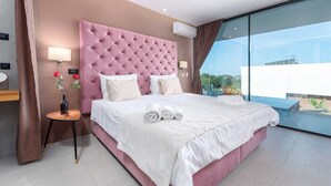 Ein modernes Hotelzimmer mit einem gepolsterten, pinkfarbenen Kopfteil, einem großen Bett mit weißer Bettwäsche, einem Nachttisch mit Lampe und einer Blumenvase sowie einem großen Fenster mit Blick auf die Stadt.