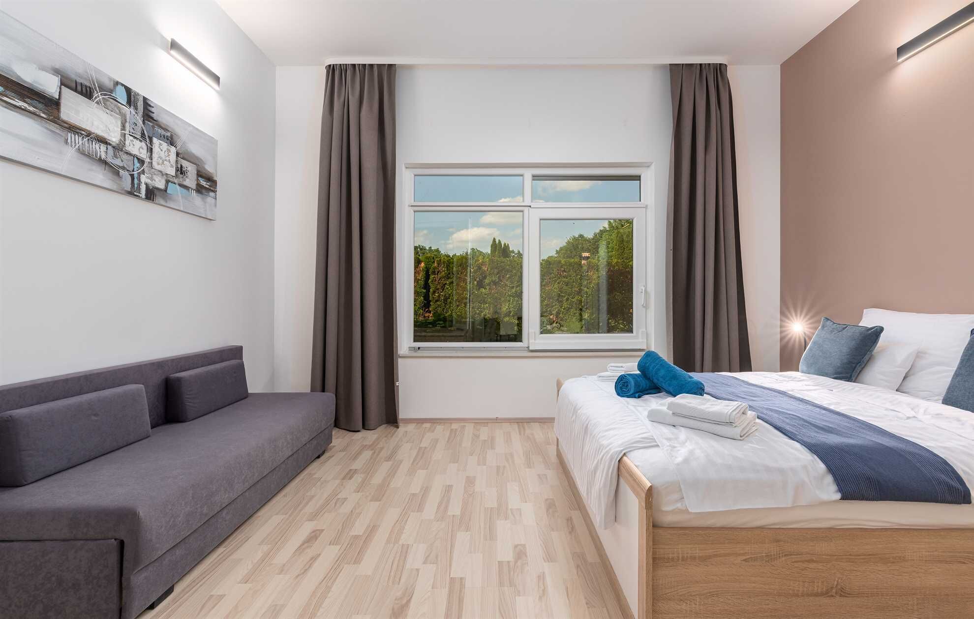 A modern hotel room with a bed, grey sofa, and a large window with curtains.