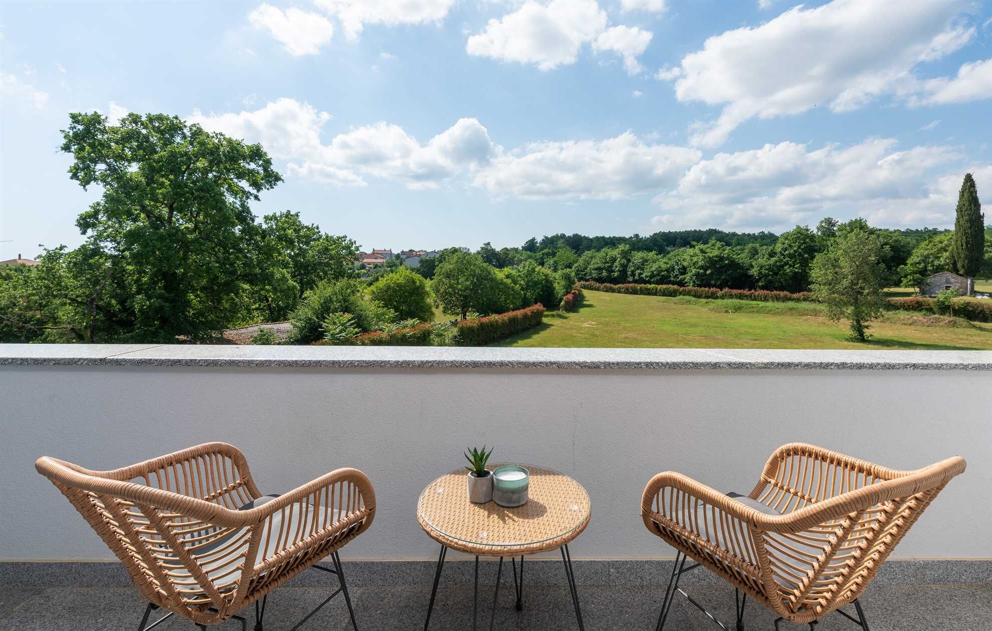 Een balkon met twee rieten stoelen en een klein rond tafeltje, met uitzicht op een aangelegde tuin en gebouwen in de verte.