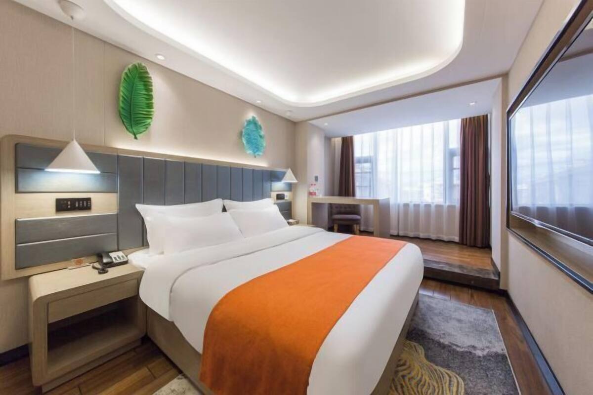 A hotel room with a bed, bedside table, lamp, and a view of the city through large windows.