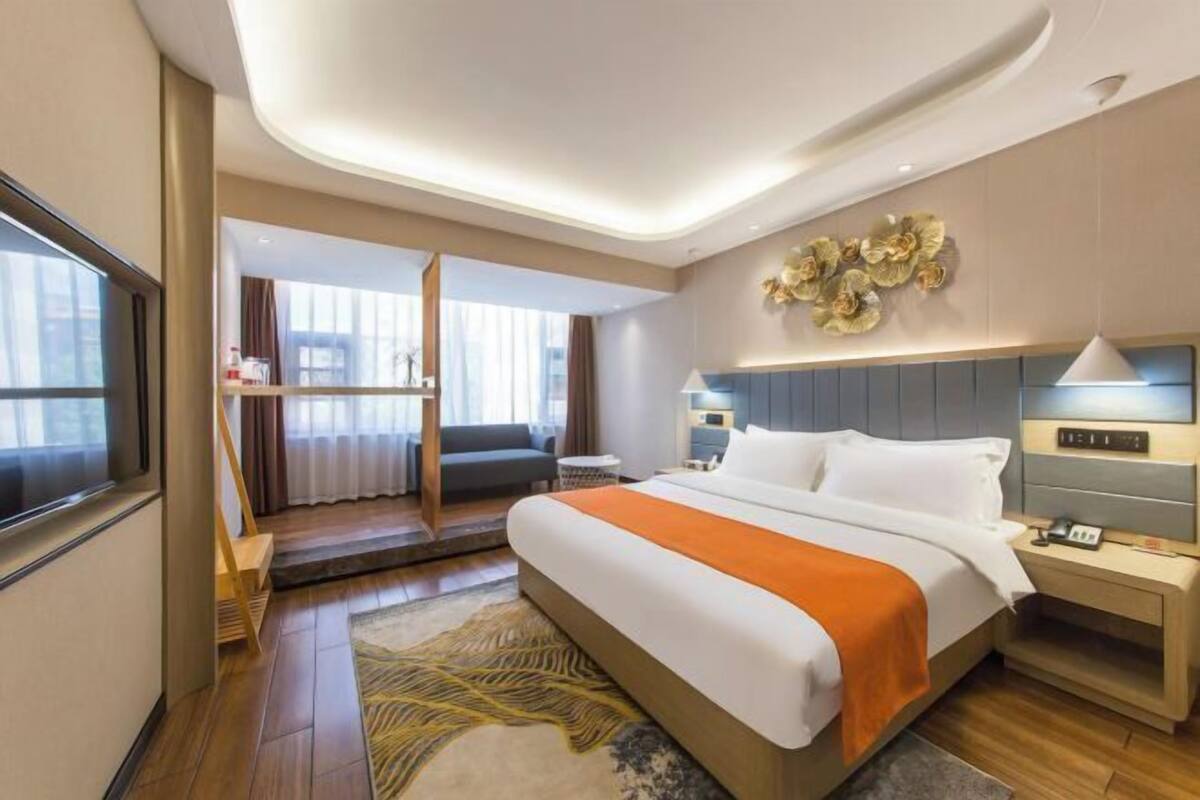 A modern hotel room with a large bed, a desk, a chair, a sofa, and a view of the city.