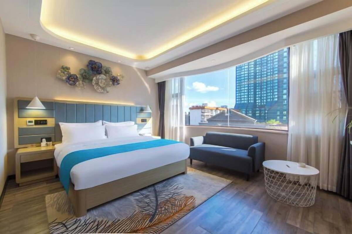 A modern hotel room with a large bed, a sofa, a round coffee table, and a view of a cityscape through large windows.
