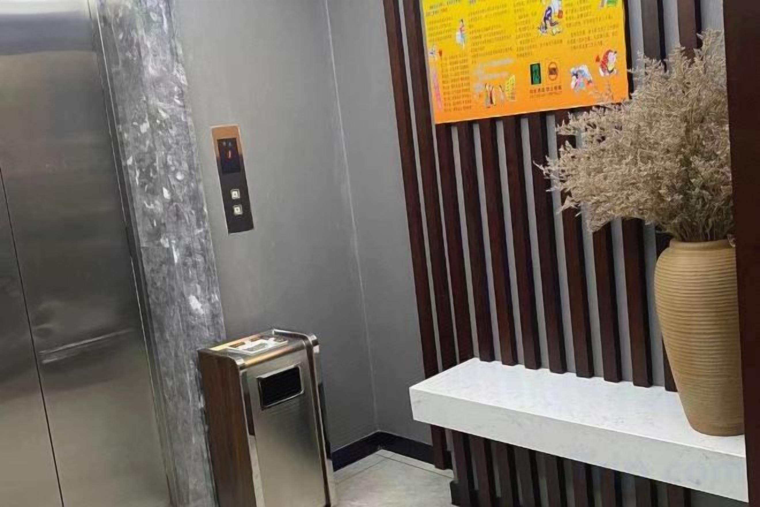 L’intérieur d’un ascenseur est agrémenté d’un revêtement métallique, d’une petite table sur laquelle est posé un vase de fleurs séchées, et d’un panneau mural à rayures verticales.