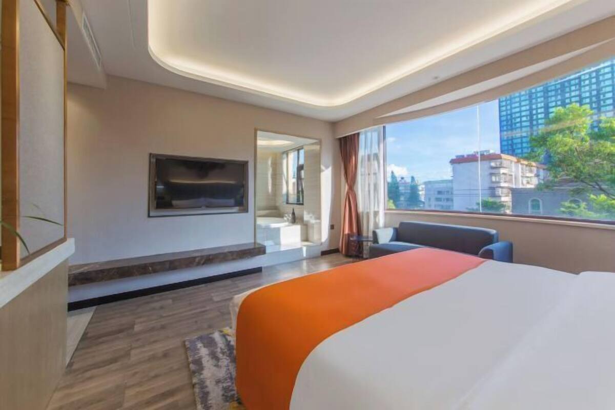A modern hotel room with a large bed, a TV mounted on the wall, a seating area with a sofa, and a view of the city through large windows.