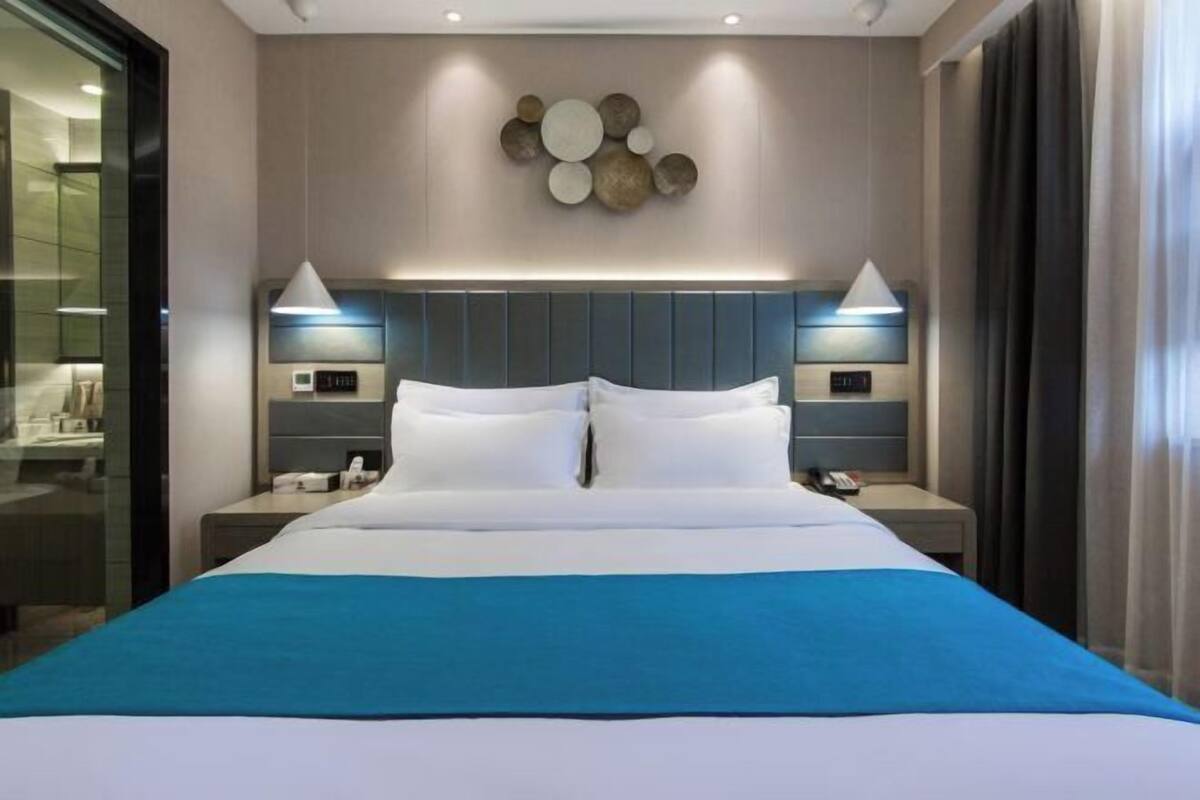A modern hotel room with a large bed, bedside tables, a wall-mounted clock, and a decorative wall piece.