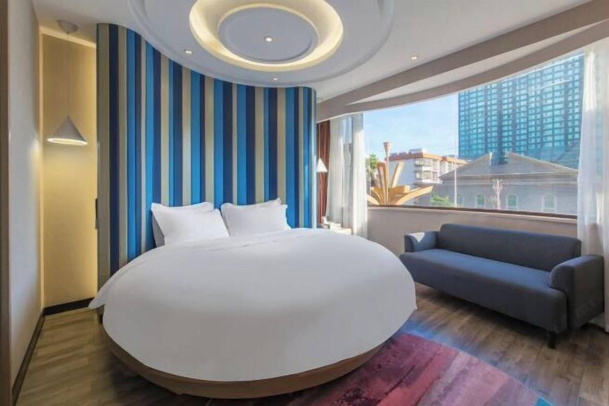 A hotel room with a large bed, a sofa, a view of a building, and a striped wall.