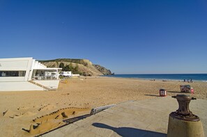 Ein Strand mit einem weißen Gebäude, Treppen davor und einem sandigen Bereich mit einigen Personen.