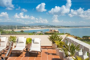 A terrace with lounge chairs and potted plants, overlooking a beach and ocean.