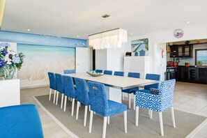 A modern dining area with a long table and blue patterned chairs, a white ceiling, and a view of the sea through a large window.