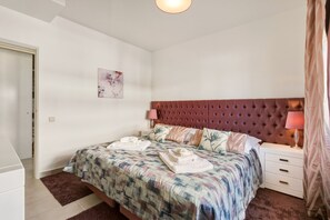 A bedroom with a bed, bedside tables, a nightstand, a lamp, a door, and a wall-mounted artwork.