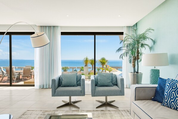 A modern living room with a view of the sea, featuring two armchairs, a sofa, a floor lamp, and a potted palm tree.