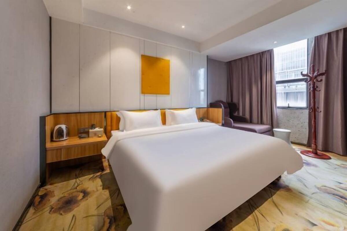 Une chambre d’hôtel avec un grand lit, une table de chevet en bois, une chaise, un porte-manteau et une fenêtre avec des rideaux.