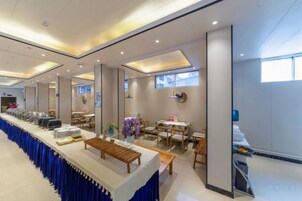 Una larga mesa de buffet con mantel azul, rodeada de sillas y mesas, en una habitación bien iluminada con techo alto y ventanas amplias.