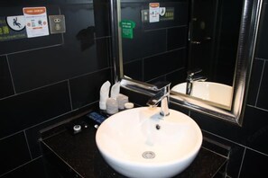A bathroom with a white sink, a mirror, and black tiled walls. There are toiletries on the counter and a green sign on the wall. - Huanyu Hotel (Lingshan Bus Terminal) (Lingshan)