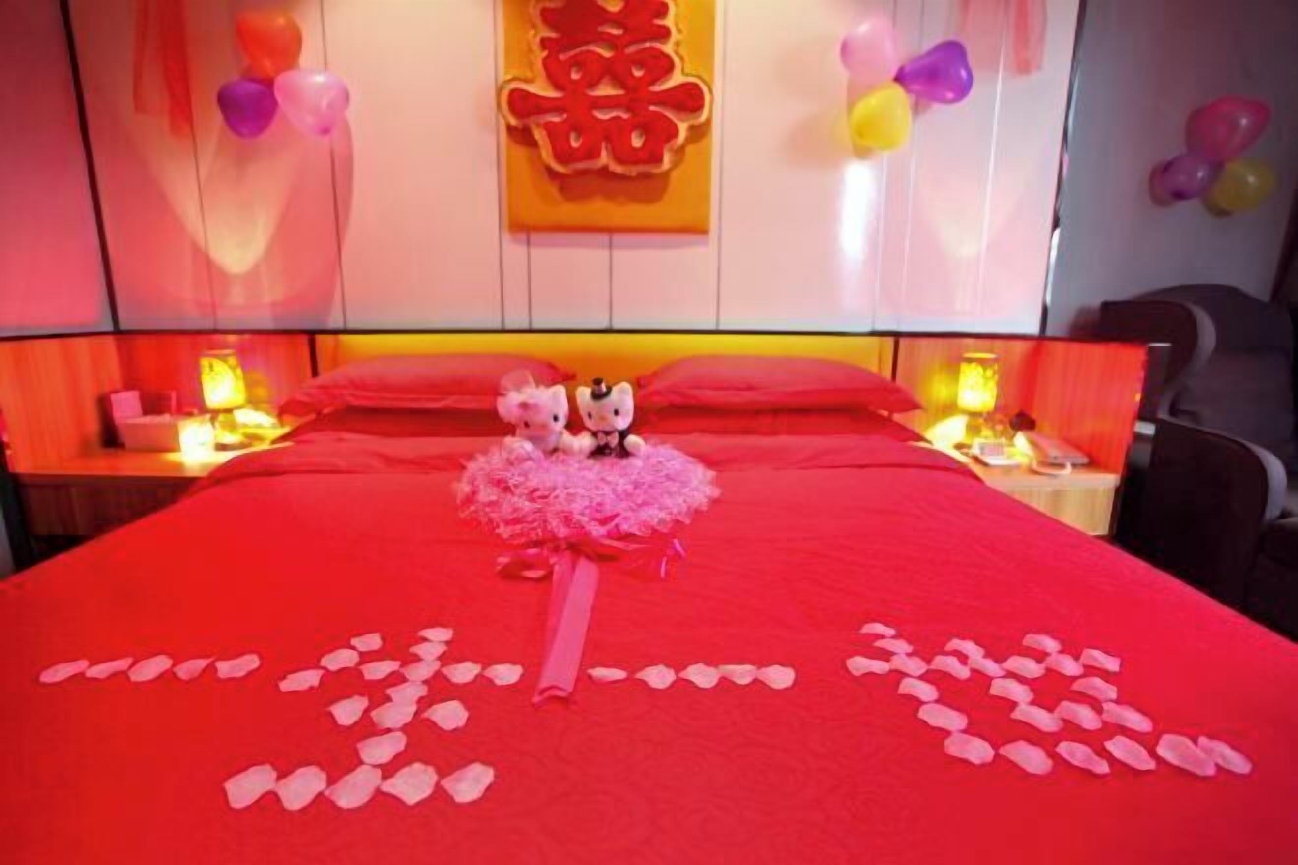 A hotel room with a red bedspread, a decorative heart-shaped arrangement on the bed, and a wall with Chinese characters.