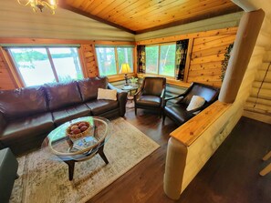 A cozy living room with a brown leather sofa, two armchairs, a glass coffee table with magazines, and a wooden bench. Large windows offer a view of trees and a lake. - Peaceful & Private Log Cabin Lake House (Brimson)