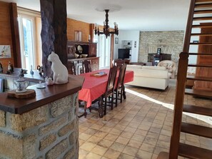 A cozy living room with a fireplace, a dining area with a red tablecloth, and a kitchen area with stone walls. - Maison typique pour 6 personnes, jardin clos et fleuri, proche plage (Treffiagat)