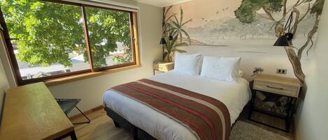 A bedroom with a bed, a desk, a window with a view of trees, a nightstand, and a wall mural.