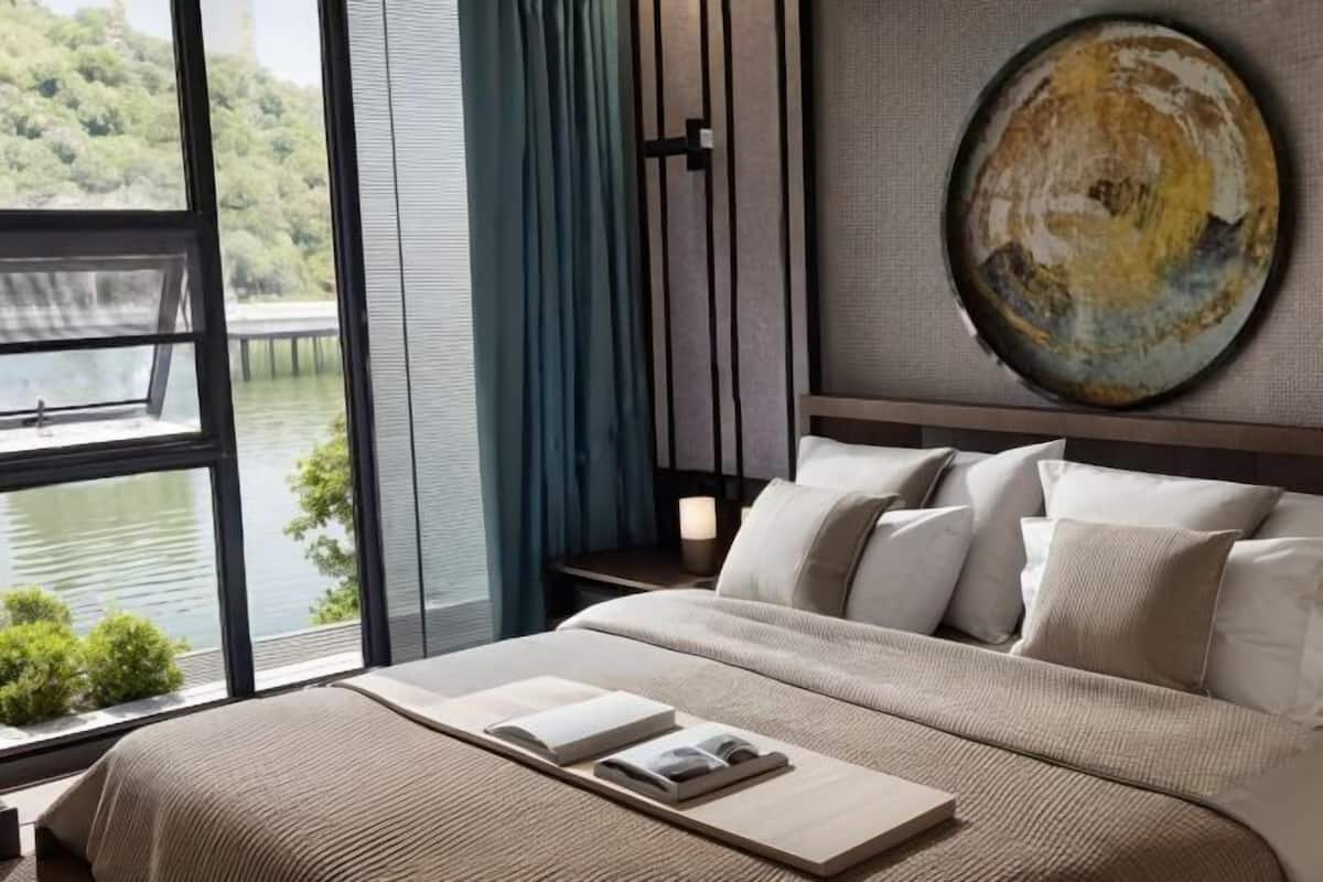 A bedroom with a large window overlooking a body of water, a bed with a view, and a round painting on the wall.