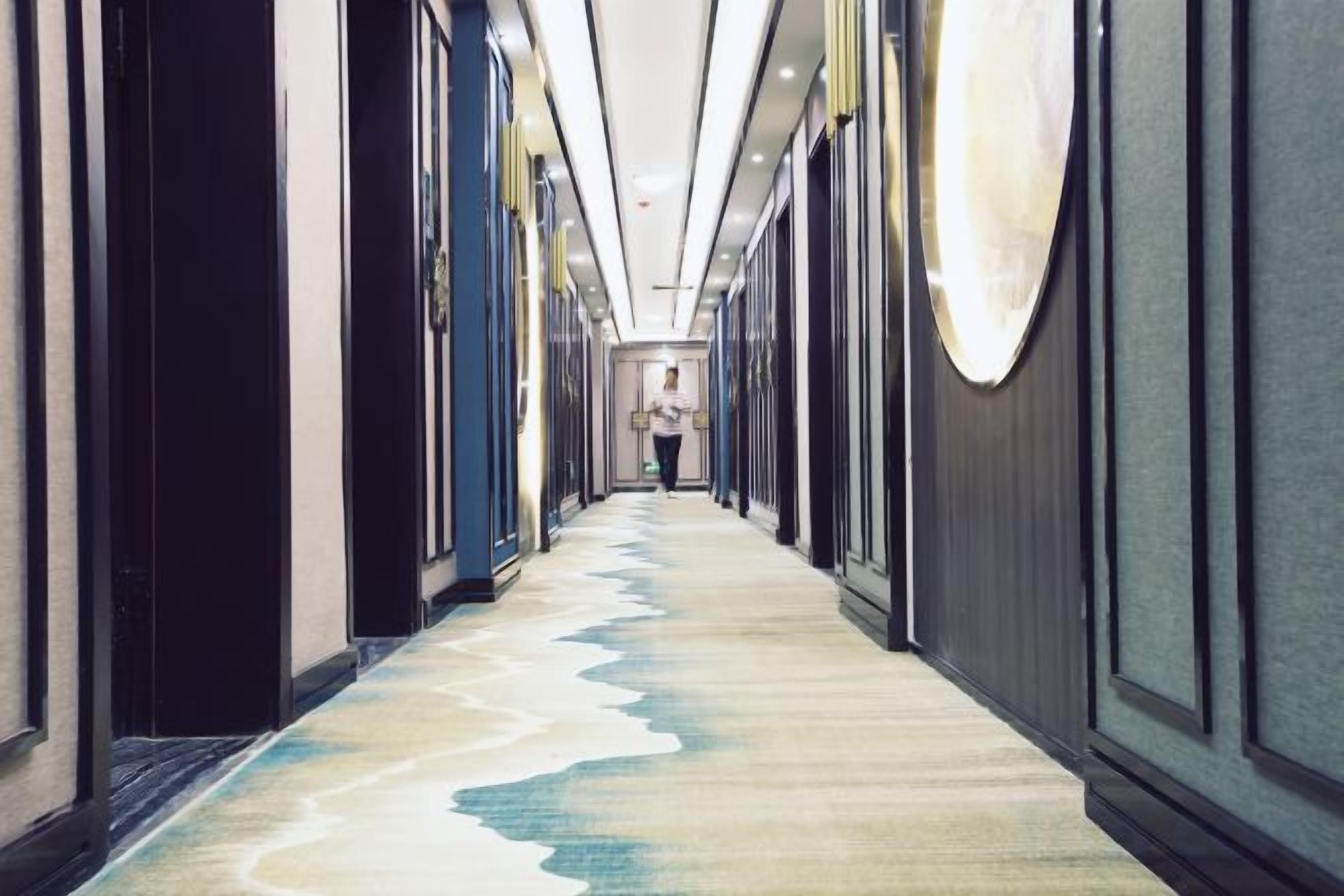 A hotel corridor with a carpeted floor, doors on both sides, and a person walking away from the camera.