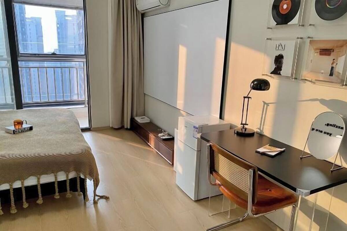 A modern bedroom with a bed, a desk, a chair, a small refrigerator, and a window with a view of buildings.