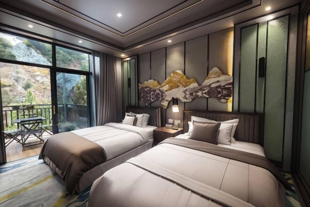 A hotel room with two beds, a large window with a view of a mountainous landscape, and a small balcony with a table and chairs.