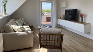 A modern living room with a sofa, a wicker chair, a TV, and a small table. - Modern holiday apartment for 2-4 people, free WLAN, summer/winter offers (Norden)