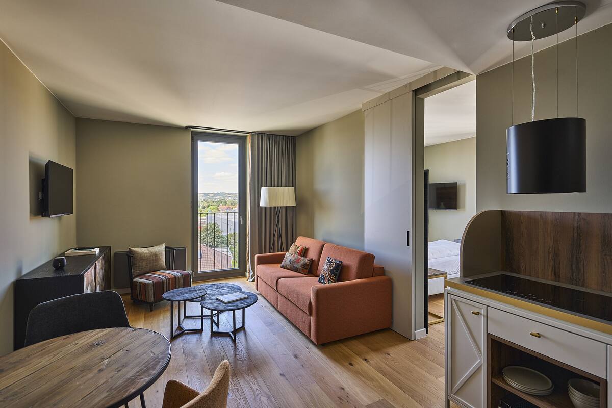 A modern hotel room with a sofa, dining table, and kitchenette.