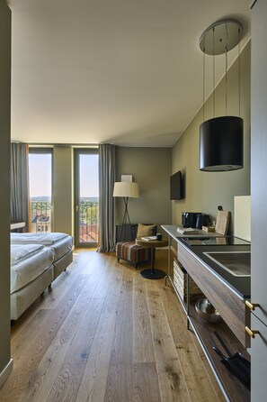 Habitación de hotel con balcón, cama, escritorio con lámpara, televisor y zona de cocina.