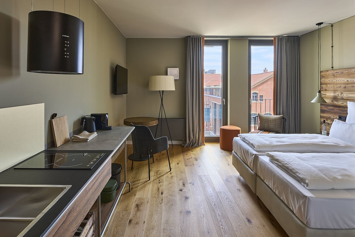 A hotel room with a bed, a desk with a chair, a standing lamp, and a view of buildings through a window.
