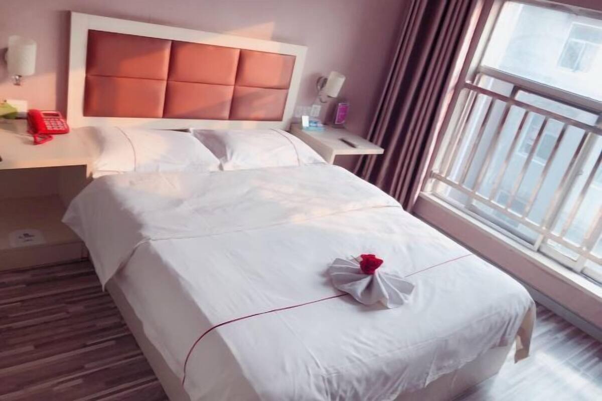 Una habitación de hotel con una cama, una mesita de noche con teléfono, una ventana con cortinas y un cabecero.