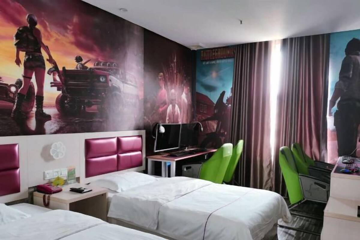 Habitación de hotel con dos camas, un escritorio con computadora, sillas verdes y un mural grande de un videojuego.
