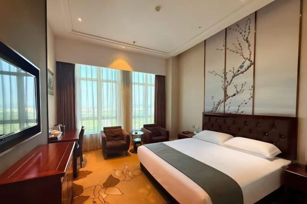 A hotel room with a large bed, a desk, two armchairs, a small table, and a large window.
