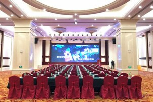 Un salón de conferencias con filas de mesas y sillas, una pantalla grande y un escenario.