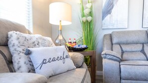 Ein Wohnzimmer mit einem grauen Sofa, einem grauen Sessel, einer Lampe, einem kleinen Tisch mit einer Obstschale und einer Pflanze.
