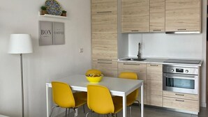 A modern kitchen with wooden cabinets, a white countertop, and a built-in oven. There is a dining table with yellow chairs and a bowl of fruit.