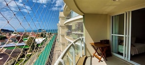 Un balcón con vista a la ciudad y al océano, una mesita pequeña y una cama con ventana.