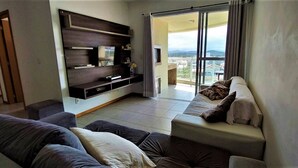 A living room with a sofa, a TV mounted on a wooden panel, and a balcony with a view. - Nautilus Caminho do Sol em Penha-SC (Penha)