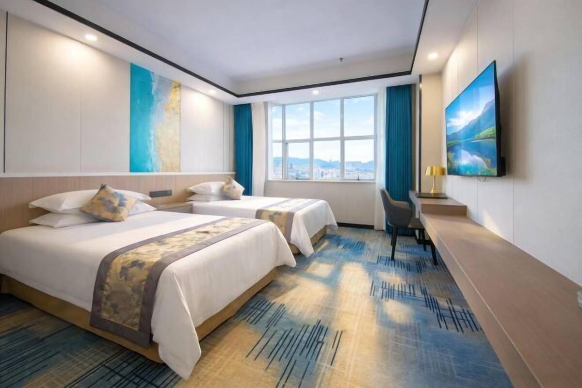 A hotel room with two beds, a desk, a chair, a large window, and a painting on the wall.