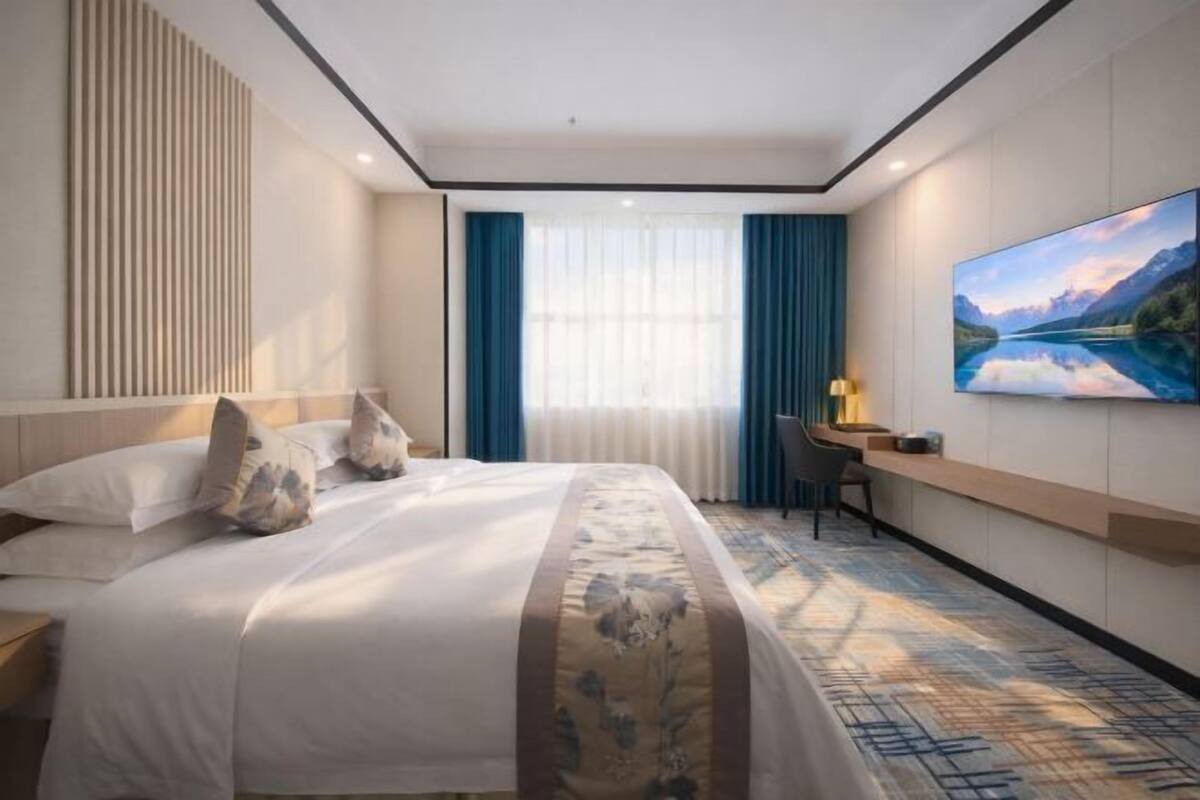 A hotel room with a bed, a desk, a chair, a large window with curtains, and a painting on the wall.