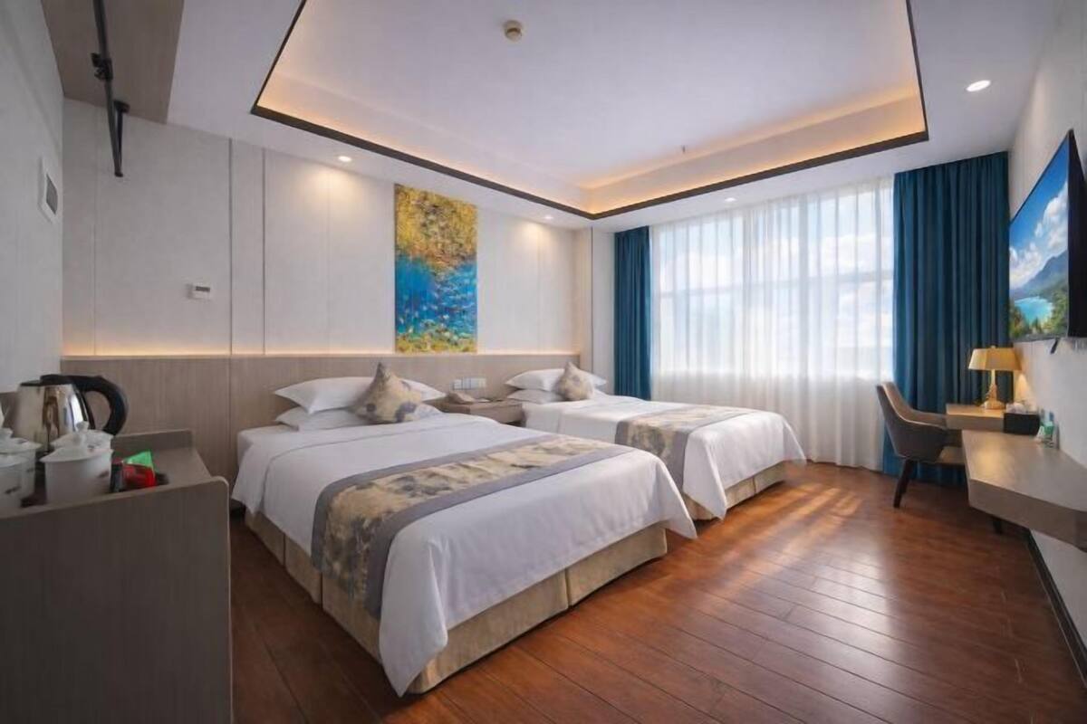 A hotel room with two beds, a desk, a chair, a painting on the wall, and a window with curtains.