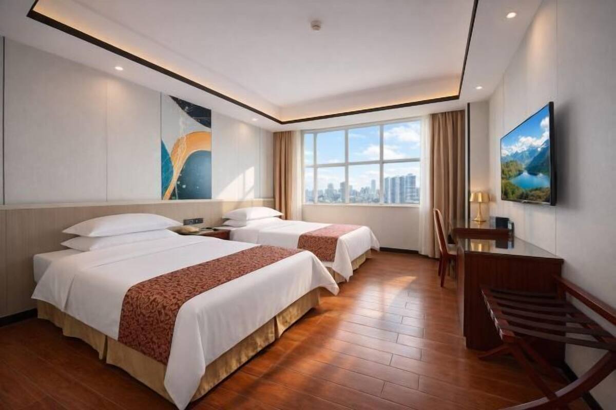 A hotel room with two beds, a desk, a chair, a TV, and a large window with a city view.