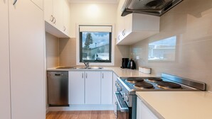 Une cuisine moderne avec des meubles blancs, un lave-vaisselle, une cuisinière et une fenêtre donnant sur un arbre et un immeuble.